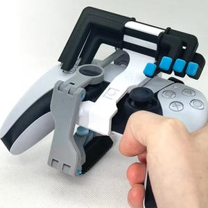 May include: A white and black gaming controller with a grey and black attachment, held in a hand. The attachment has blue accents and a black frame. The controller has a black joystick and grey buttons.