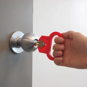 以下が含まれることがあります： 赤いキーターナーが銀色のドアノブに差し込まれています。キーターナーには握りやすい大きなハンドルが付いています。ドアノブは灰色のドアに取り付けられています。背景は白です。