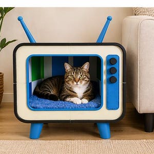 May include: A tabby cat is nestled in a novelty cat bed designed to resemble a vintage television set. The bed features a cream-coloured frame with blue detailing, including the legs, aerials, and control panel. The cat rests on a blue cushion.