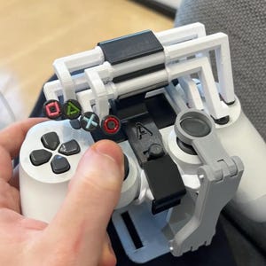 Op de afbeelding: Een witte en zwarte gamecontroller met een op maat gemaakte 3D-geprinte accessoire. De accessoire heeft een wit frame, een zwarte clip en een grijze steunarm. De controller heeft gekleurde knopsymbolen en zwarte richtingknoppen.