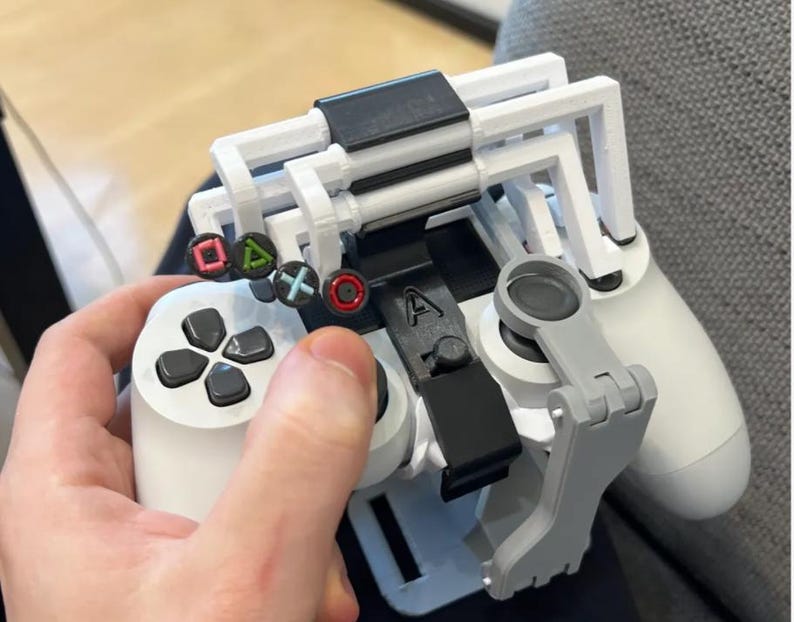 Op de afbeelding: Een witte en zwarte gamecontroller met een op maat gemaakte 3D-geprinte accessoire. De accessoire heeft een wit frame, een zwarte clip en een grijze steunarm. De controller heeft gekleurde knopsymbolen en zwarte richtingknoppen.