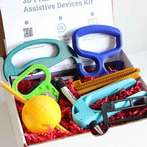 May include: A box filled with 3D-printed assistive devices in various colors, including blue, green, yellow, and black. The kit contains tools like a saw, a handle, and a writing aid, all nestled in red paper. The box has text that reads "3D Printed Assistive Devices Kit."