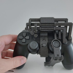 May include: Black video game controller with a gray attachment. The controller has a directional pad, buttons, and analog sticks. The gray attachment has a complex, geometric design, and is attached to the top and bottom of the controller.