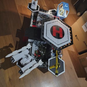 Può includere: Un set di gioco base militare a più livelli con un grande eliporto esagonale con un simbolo H rosso, un accessorio lanciamissili bianco, una gru gialla e una torre di osservazione blu contenente una piccola figura, tutto in plastica.