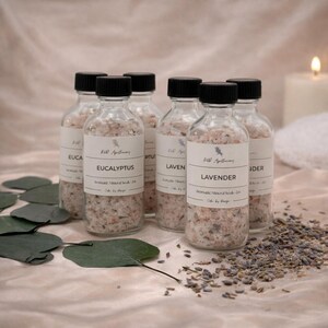 May include: Six clear glass bottles with black caps, filled with mineral salts. Each bottle has a label with the text "Eucalyptus" or "Lavender". The bottles are surrounded by eucalyptus leaves and lavender buds. A lit candle is in the background.