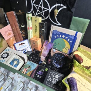 May include: A collection of items including a black tote bag with a white pentagram, a crystal pendulum, incense sticks, a tarot mug, a notebook with a moon illustration, various small boxes, tarot cards, enamel earrings, and loose gemstones like amethyst and tiger's eye.