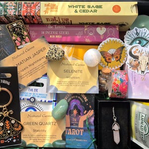 May include: A gift box containing various items including a moth keyring, incense sticks labelled White Sage & Cedar and Red Rose, a deck of tarot cards, a crystal pendant, loose gemstones like Dalmatian Jasper and Selenite, and tea.