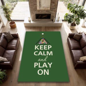 May include: A green rectangular rug with the words "KEEP CALM and PLAY ON" in white. Above the text is a triangle of billiard balls and two crossed pool cues. The rug is in a living room with two brown leather sofas.