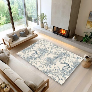 May include: A rectangular rug with a cream backdrop and a blue toile design depicting deer and foliage. The rug is in a living room with a fireplace and two sofas. The rug's design is reminiscent of classic French decor.