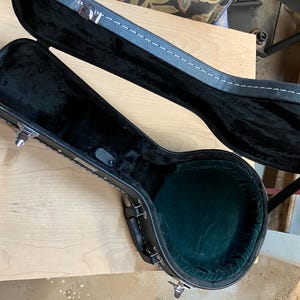 May include: An open black banjo case with a green velvet interior. The case is made of black leather with white stitching and silver latches. The case is on a light wood surface.