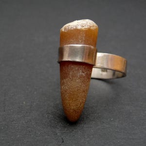 Könnte beinhalten: Ein einzigartiger Ring mit einem natürlichen Bernstein, der in einem Silberband gefasst ist. Der Bernstein hat eine warme, braune Farbe und eine strukturierte Oberfläche. Das Band des Rings ist aus poliertem Silber.