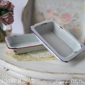 May include: Two small, rectangular, white enamelware pans with red and blue trim. The pans have a distressed, vintage look. The pans are on a white surface with a lace doily.