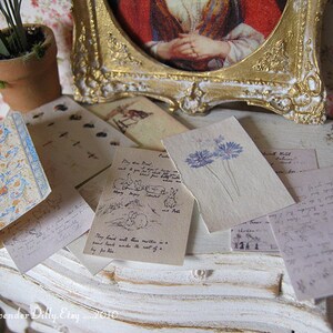 May include: A collection of vintage-style paper ephemera with floral and botanical illustrations. The papers are arranged on a white painted surface with a gold-painted frame in the background.