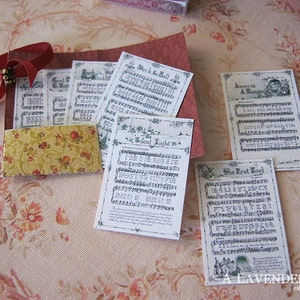 May include: A collection of vintage-style paper ephemera featuring sheet music and decorative elements. The pieces include song titles like "Silent Night" and "The First Noel", with a burgundy ribbon and a small floral patterned card.