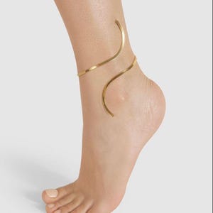 May include: A gold-colored ankle bracelet wraps around the ankle and foot. The jewelry has a minimalist design, with a curved, open shape. The bracelet is made of a thin, metallic material and is worn on a light-skinned foot.