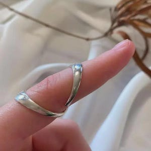 May include: A silver ring with a unique, open design. The ring curves around the finger, with a smooth, polished finish. The ring is worn on a finger, showcasing its elegant shape and reflective surface. The ring's design is modern and minimalist.