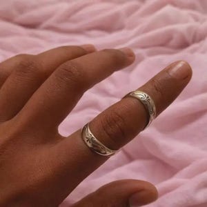 May include: Two silver rings with ornate floral designs. The rings are worn on a finger, showcasing their intricate details. The rings have a polished finish, reflecting light. The background is a soft pink fabric.