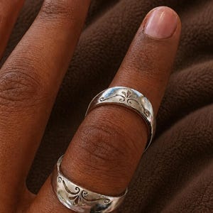 May include: Two silver rings with a floral design are displayed on a finger. The rings have a curved band and a detailed pattern. The background is a textured brown fabric.