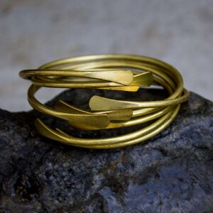 May include: A stack of gold-colored bracelets with a leaf-shaped design. The bracelets are coiled and rest on a dark, textured surface. The bracelets have a shiny, metallic finish.