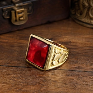 May include: A gold-toned ring featuring a large, square-shaped, deep red stone set in the center. The band of the ring displays a textured, irregular pebble-like pattern across its surface, providing a distinct contrast to the smooth, flat red stone.