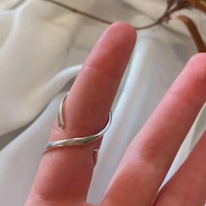 May include: A silver ring with a unique, open design. The ring wraps around the finger with a curved, flowing shape. The metal has a polished, reflective surface, creating a modern and minimalist aesthetic. The ring is worn on a finger.