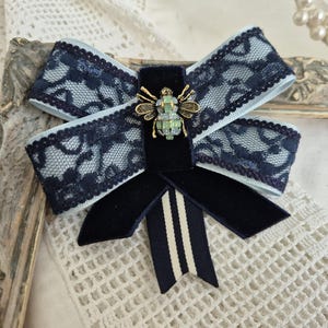 May include: A bow brooch featuring a light blue ribbon overlaid with dark navy blue lace. The centre is adorned with a metallic bee embellishment with green crystal accents. Below the bow, two dark navy velvet tails and a striped ribbon tail hang.