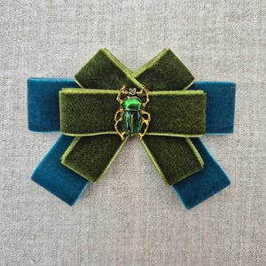 May include: A layered bow accessory crafted from velvet ribbon in olive green and teal blue. The centre of the bow features a metallic gold-toned beetle brooch with a vibrant, shiny green enamel finish on its shell, creating a distinct focal point.