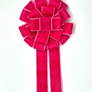 Könnte beinhalten: Eine leuchtend fuchsiafarbene Samtschleife mit einem geschichteten Design. Die Schleife hat einen zentralen quadratischen Knoten, umgeben von überlappenden Quadraten und zwei langen, rechteckigen Bändern. Die leuchtend rosa Farbe und die weiche Textur sind auffällig.