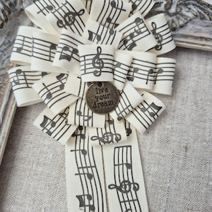 Pode incluir: Uma roseta decorativa feita de fita na cor creme estampada com notas musicais e pautas pretas. O centro apresenta um pingente redondo de metal na cor bronze antigo com o texto "live your dream" e duas fitas longas.