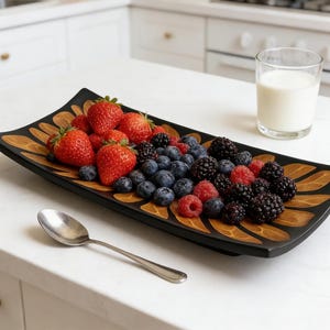 May include: A rectangular black serving tray with a decorative leaf design holds a colorful assortment of fresh berries. Strawberries, blueberries, raspberries, and blackberries are arranged on the tray. A glass of milk and a spoon are also present.
