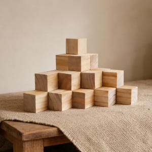 May include: A pyramid of light-colored wooden blocks sits on a burlap-covered table. The blocks are arranged in a stepped pattern, with each layer decreasing in size. The background features neutral tones and decorative vases.