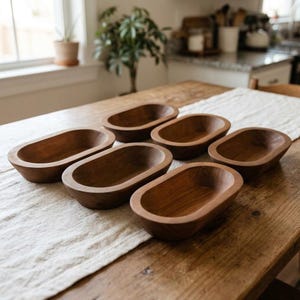 May include: A set of six small, oval-shaped wooden bowls with a smooth, dark brown finish. Each bowl features a deep, rounded interior and a thick, solid rim, arranged in a cluster on a textured, light-colored fabric runner.