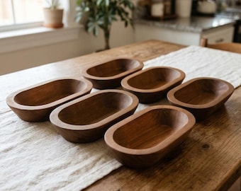 May include: A set of six small, oval-shaped wooden bowls with a smooth, dark brown finish. Each bowl features a deep, rounded interior and a thick, solid rim, arranged in a cluster on a textured, light-colored fabric runner.