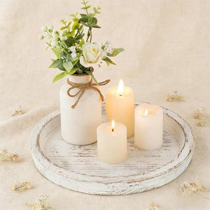 May include: A white, distressed wooden tray holds a white vase with a twine bow, a floral arrangement, and three lit pillar candles. The candles are different sizes and have a warm glow, creating a cozy ambiance.