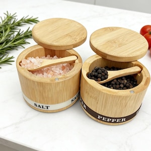 May include: Two bamboo salt and pepper containers with wooden lids and small spoons. One container holds pink salt, the other black peppercorns. The containers are labeled "SALT" and "PEPPER". Fresh rosemary sprigs and tomatoes are in the background.