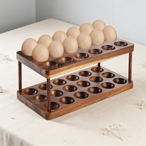 May include: A two-tiered wooden egg holder with a dark brown finish. The top tier holds a dozen brown eggs, while the bottom tier has space for another dozen. The holder is made of solid wood and has a simple, elegant design.