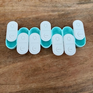 May include: A light blue weekly pill organizer with seven white compartments. Each compartment is labeled with a day of the week and features a sun or moon icon. The organizer is designed to hold and dispense medication.