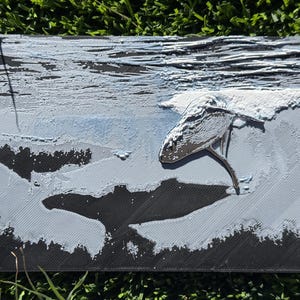 May include: A textured artwork showing a whale breaching the ocean's surface. The whale is silver against blue and grey tones. The piece has a 3D effect. The artwork is rectangular.