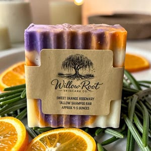 Op de afbeelding: Een rechthoekige zeep met lagen paars, oranje en wit. Een bruin etiket met de tekst "Willow Root Skincare Co." en "Sweet Orange Rosemary Tallow Shampoo Bar ca. 113-142 gram" is om de zeep gewikkeld. Sinaasappelschijfjes en rozemarijn omringen de zeep.