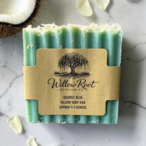 Puede incluir: Una pastilla de jabón de sebo azul coco rectangular con una etiqueta de papel kraft. La etiqueta dice "Willow Root Skincare Co." y "Coconut Blue Tallow Soap Bar Aprox. 113-142 gramos." El jabón es azul claro con copos blancos. Una mitad de coco está en el fondo.