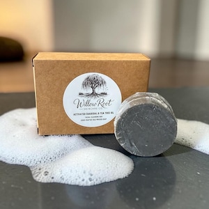 May include: A round, dark gray facial cleansing bar wrapped in clear plastic, positioned next to a rectangular brown cardboard box with a white circular label featuring a willow tree illustration and the text Willow Root Skincare Co.