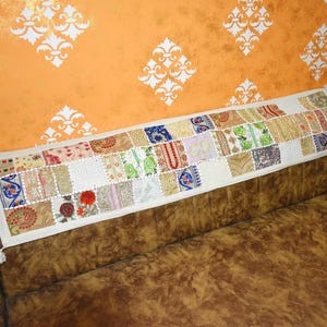 May include: A long, rectangular fabric runner featuring a patchwork design of various embroidered squares in colors including red, blue, green, and gold on a cream-colored base. The runner has small white tassels at each of the two ends.