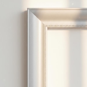 Puede incluir: Primer plano de un marco de fotos plateado con una superficie texturizada y un borde interior con cuentas. El marco está colocado contra una pared blanca, con una iluminación suave que crea sombras.