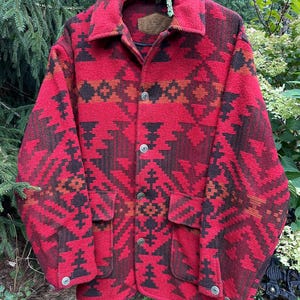May include: A long-sleeve button-up jacket featuring a bold red, dark brown, and orange geometric pattern. The jacket includes a pointed collar, two front pockets with flaps, and metallic buttons. A brown leather-like brand patch is visible inside.