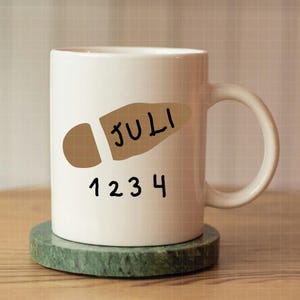 May include: White ceramic mug with a brown footprint design and the word "JULI" and the numbers "1234" in black. The mug sits on a green marble coaster. The mug has a curved handle.
