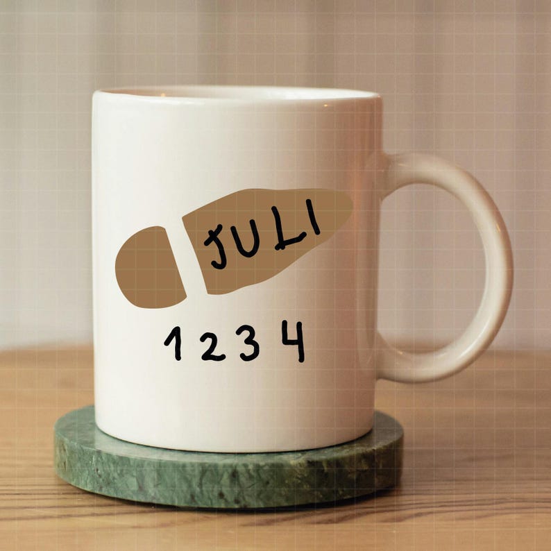 May include: White ceramic mug with a brown footprint design and the word "JULI" and the numbers "1234" in black. The mug sits on a green marble coaster. The mug has a curved handle.