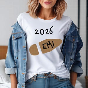 May include: White t-shirt with the year "2026" in black script above a brown footprint graphic with the letters "EMI" inside. The shirt is worn with a denim jacket and blue jeans.