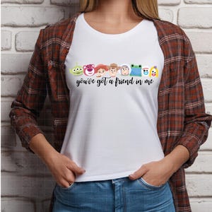 Może przedstawiać: Biały t-shirt z kr&oacute;tkim rękawem z grafiką przedstawiającą r&oacute;żne postacie z kresk&oacute;wek w rzędzie nad czarnym napisem kursywą "you've got a friend in me". Koszulka jest założona pod brązowo-czarną koszulę w kratę z długim rękawem.