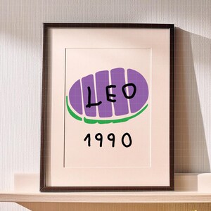 May include: Framed art print featuring a purple oval design with the word "LEO" in black letters, and the number "1990" below. The design is set against a white background, framed in a dark brown frame.
