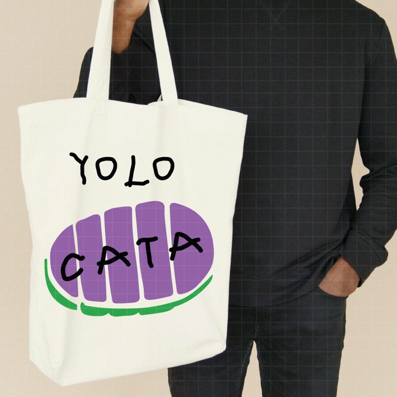 May include: A white tote bag with the words "YOLO CATA" in black and purple lettering. The word "CATA" is inside a purple oval with green trim. The bag has white handles.